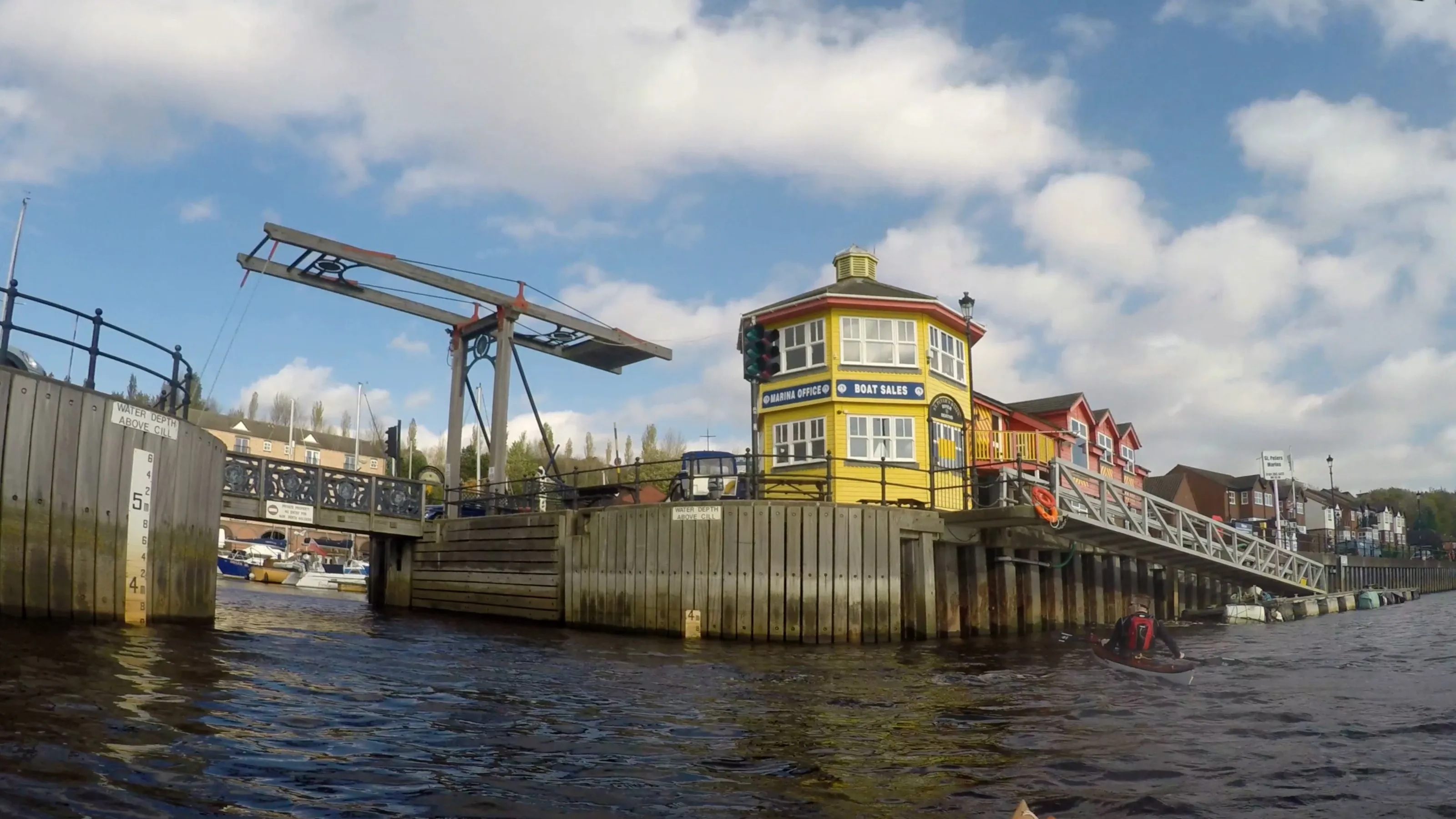 The entrance to St Peters Marina seen from the River Tyne: the yellow marina office, the raised lifting footbridge and the water depth board